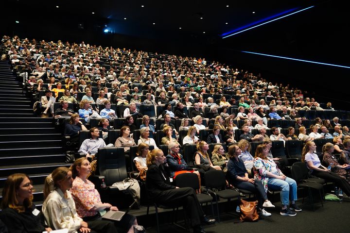 Billeet viser et fyldt auditorie, hvor alle sæder på 15 rækker er besat af mennesker, der er mødt op i hverdagstøj og har deres fulde opmærksomhed rettet mod en taler, der ikke er med på billedet.