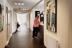 Billedet viser en lang gang i en lægeklinik, hvor en person står lænet ind over en skranke og taler med en medarbejder, der ikek kan ses. Personen på gangen ses bagfra.