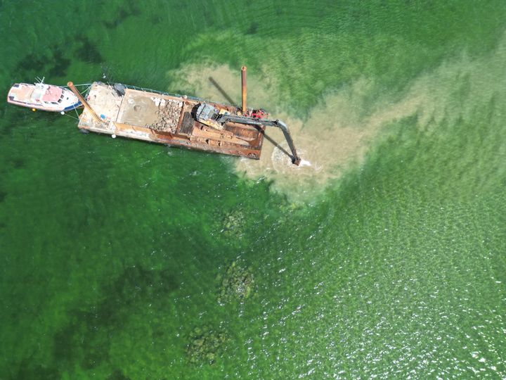 Luftfoto af pram med gravemaskine, der etablerer et stenrev i havet.