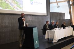 En mand holder en tale ved et podie foran et panel og publikum.