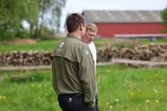 To mennesker står og taler i en grøn mark med en rød lade i baggrunden.