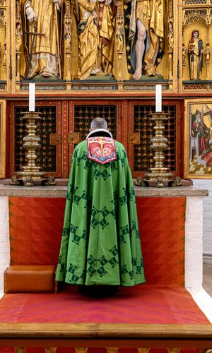 Biskoppen i Aarhus Domkirke iført bispekåbe, designet af H.M. Dronning Margrethe i 1999. På kåben har Dronning Margrethe broderet symbolerne for tro, håb og kærlighed i form af kors, anker og hjerte. Øverst ses bogstaverne alfa og omega fra det græske alfabet, der symboliserer verdens begyndelse og ende.
