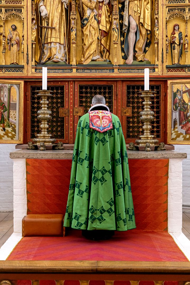 Biskoppen i Aarhus Domkirke iført bispekåbe, designet af H.M. Dronning Margrethe i 1999. På kåben har Dronning Margrethe broderet symbolerne for tro, håb og kærlighed i form af kors, anker og hjerte. Øverst ses bogstaverne alfa og omega fra det græske alfabet, der symboliserer verdens begyndelse og ende.