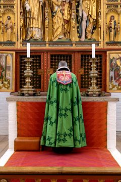 Biskoppen i Aarhus Domkirke iført bispekåbe, designet af H.M. Dronning Margrethe i 1999. På kåben har Dronning Margrethe broderet symbolerne for tro, håb og kærlighed i form af kors, anker og hjerte. Øverst ses bogstaverne alfa og omega fra det græske alfabet, der symboliserer verdens begyndelse og ende.