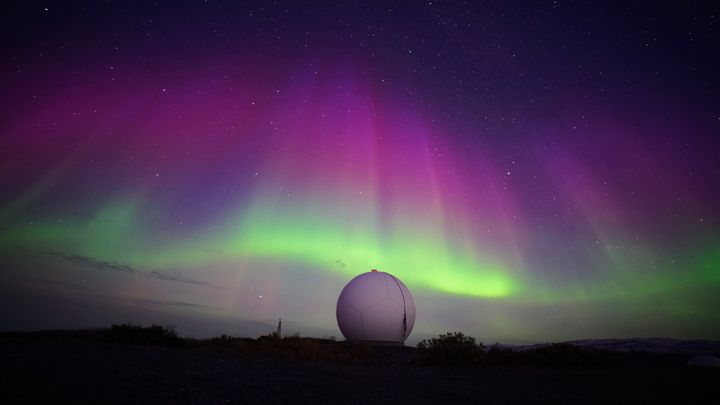 Nattefoto af stort, rundt og hvidt teleskop under en himmel med nordlys