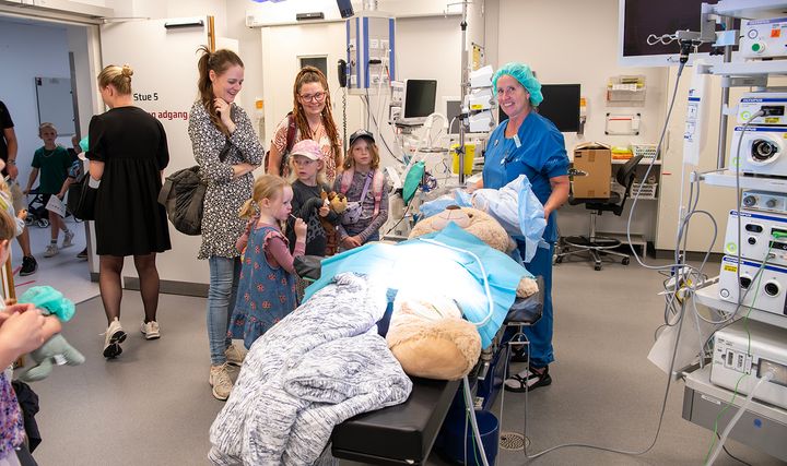 En bamse ligger på et operationsleje, børn og voksne står og kigger på til Åbent Hospital i Randers.
