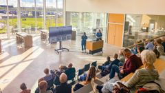 To personer i blå kittel holder oplæg for deltagere i stort lyst lokale.