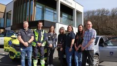 Syv personer står udenfor et sundhedshus i solskin mellem en gulgrøn akutbil og en grå bil fra Lemvig Kommune. Gruppen står foran bygningen, og i baggrunden ses store vinduer og træer.