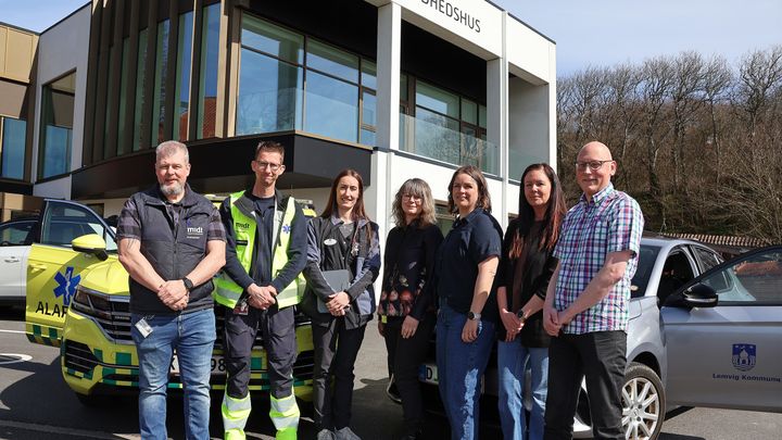Syv personer står udenfor et sundhedshus i solskin mellem en gulgrøn akutbil og en grå bil fra Lemvig Kommune. Gruppen står foran bygningen, og i baggrunden ses store vinduer og træer.