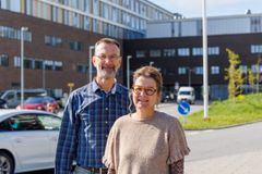 Portræt af mand i blå skjorte og kvinde i lysbrun bluse med et hospital i baggrunden.