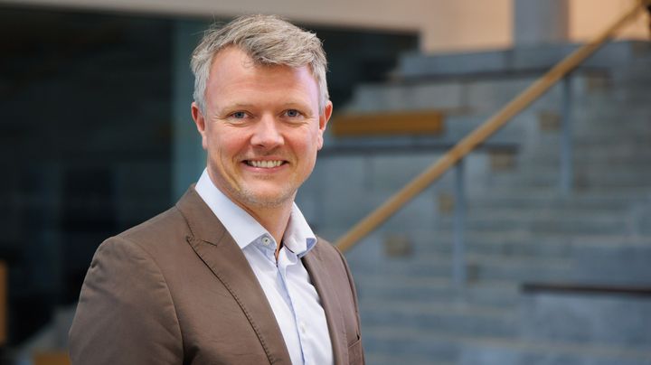 Portræt af mand i hvid skjorte og brun jakke foran grå trappetrin i baggrunden.