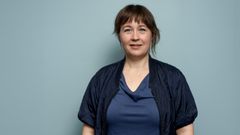 Portrætfoto af kvinde i blå hospitalsuniform på en grå baggrund.