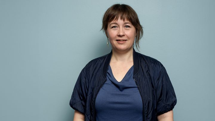 Portrætfoto af kvinde i blå hospitalsuniform på en grå baggrund.