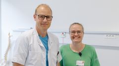 To personer i hospitalstøj står i et klinisk miljø og smiler til kameraet.