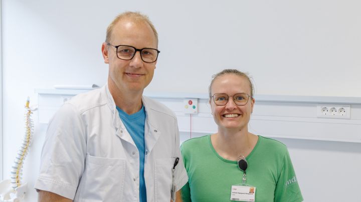 To personer i hospitalstøj står i et klinisk miljø og smiler til kameraet.