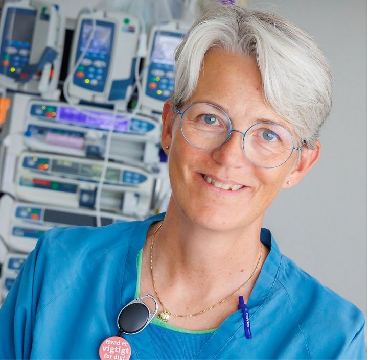 Anne Højager Nielsen i blå sygehusuniform smiler foran medicinsk udstyr på et intensivafsnit.