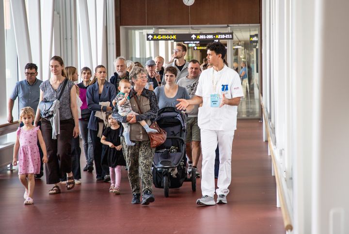 En gruppe mennesker - børn, kvinder og mænd - går på en lang hospitalsgang. De vises rundt af en medarbejder i hvidt hospitalstøj.