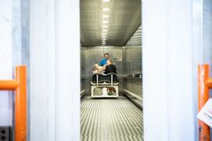 Kvinde og to børn sidder på hospitalskøretøj i elevator, smiler til kameraet, mens elevatordørene er ved at lukke i.