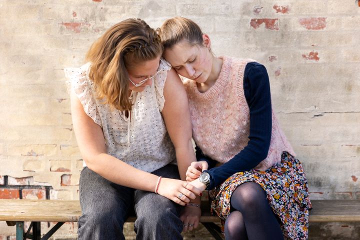 To unge piger sidder tæt sammen på en bænk og trøster hinanden