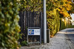 Skilt med "Til salg" ved en træindhegning langs en vej med grønne og gyldne træer.