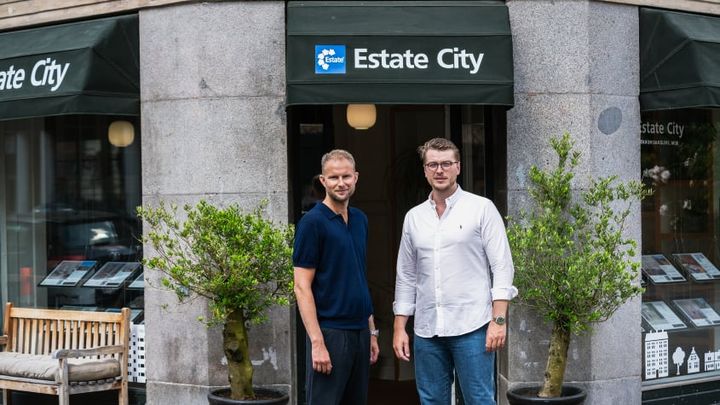To mænd står foran en ejendomsmæglervirksomhed med to planter i krukker.