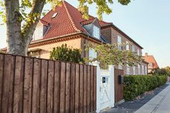 Et rødt murstenshus med rødt tegltag og hvide vinduer, omgivet af en træhegn og en hæk, set fra gaden.