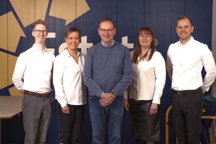 En gruppe på fem personer står foran en blå baggrund og smiler.