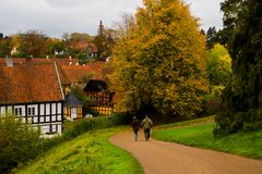 To personer går på en sti i en efterårslandskab med farverige træer og traditionelle huse i baggrunden.