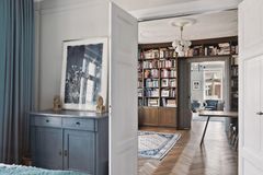 Interiør af et hjem med en åben dør til et bibliotek fyldt med bøger, en blå skænk, træmøbler og et tæppe på trægulvet.