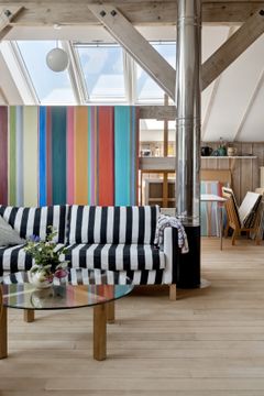 Stue med stribet sofa, glasbord, farverig stribet væg og store ovenlysvinduer.