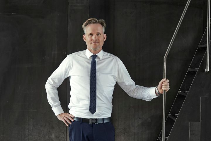 Kædechef i Ejendomsmæglerfirmaet &LIVING, Thomas Boye Jensen, står i hvid skjorte og blå slips ved en trappe.