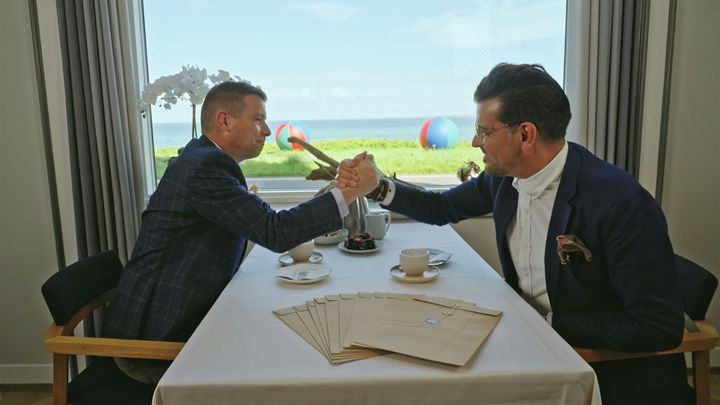 Ejendomsmæglerne Bahadir Demirhan fra Odense og Peder Kongsgaard fra Thy  giver håndtryk ved et bord med kuverter foran en havudsigt.