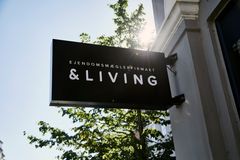Skilt ved en bygning med teksten "Ejendomsmæglerfirmaet &Living", med træer og sollys i baggrunden.