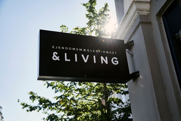 Skilt ved en bygning med teksten "Ejendomsmæglerfirmaet &Living", med træer og sollys i baggrunden.