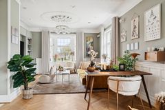 Stilfuldt indrettet stue med moderne møbler, grønne planter og kunst på væggene. Store vinduer giver naturligt lys. Nænsomt istandsat med respekt for sit ophav. Stil og arkitektur appelerer til liebhavere på boligjagt i Aalborg.