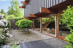 Moderne hus med store vinduer, synlige træbjælker og en have med stenbelægning og grønne buske.