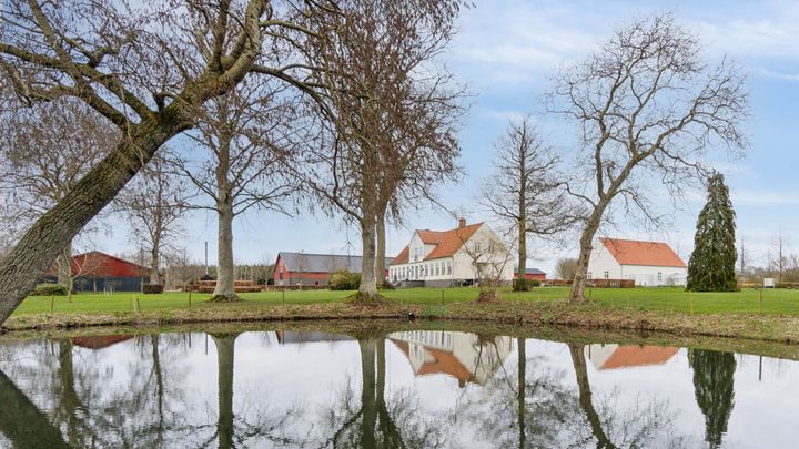 En malerisk gård ved en dam med spejlblank overflade, omgivet af nøgne træer under en klar blå himmel.