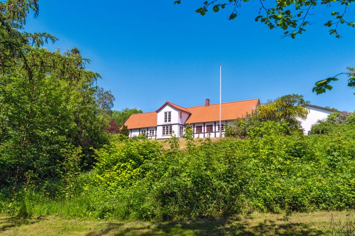 Landejendom%20p%E5%20Kalkbyvejen%20ved%20Aakirkeby%20p%E5%20Bornholm%20med%20r%F8dt%20tag%20og%20omgivet%20af%20gr%F8n%20bevoksning%20under%20bl%E5%20himmel.