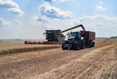 En mejetærsker og en traktor arbejder på en kornmark under en solrig himmel med skyer.