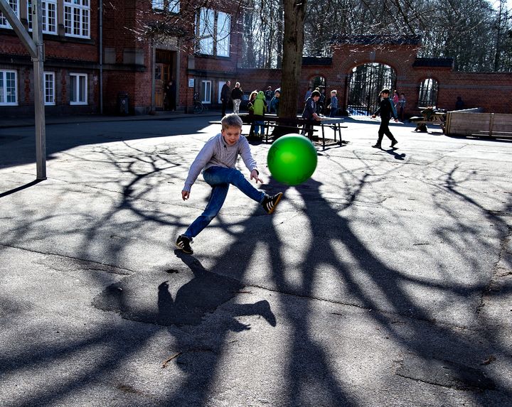 Børn i leg i en skolegård. En dreng midt i billedet sparker en fodbold i retning af fotografen.