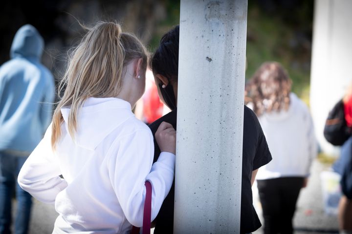 Store børn i en skolegård. To piger står helt tæt sammen med ryggen til beskueren og kigger ned i en telefon