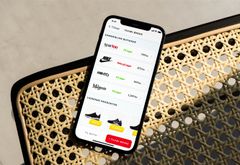 iPhone liggende på stol med et skærmbillede fra ønskeliste-appen Cupio, der viser en prissammenligning over forskellige butikker