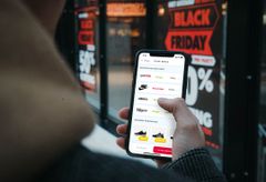 Mand holder en iPhone i hånden, der viser en prissammenligning fra ønskeliste appen Cupio. Bag telefonen ses en reklame for Black Friday.