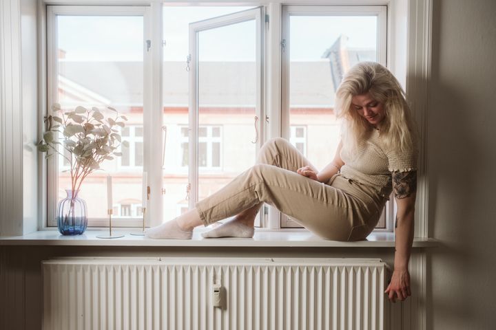 Kvinde sidder i vindueskarm og skruer på termostaten på radiatoren.
