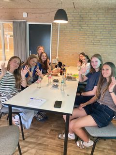 En gruppe unge piger sidder ved et bord, smilende og vinkende. De er i gang med at finde idéer til et samlingssted i Hou