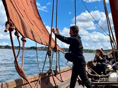 Lydkunstneren står med optageudstyr på en kopi af et vikingeskib på en fjord. Sejlet er orange.