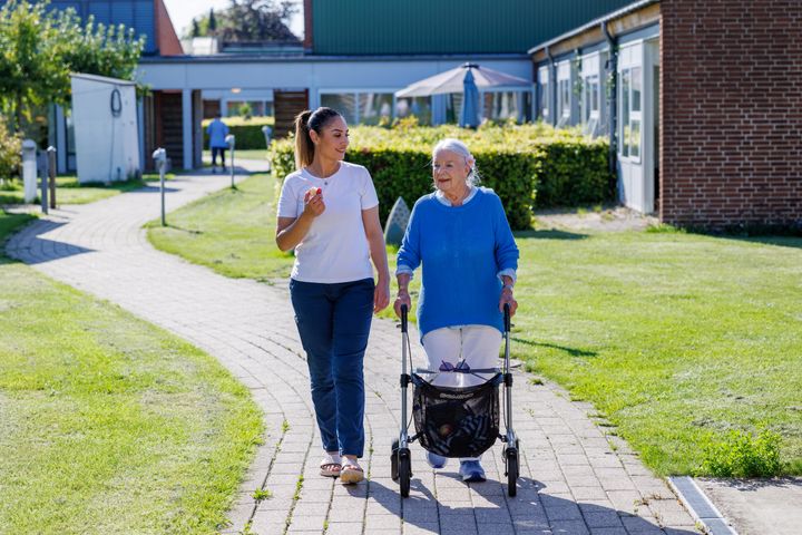 Ældre  kvinde med rolator går sammen med en yngre sosu-ansat på en sti i forbindelse med et plejehjem.