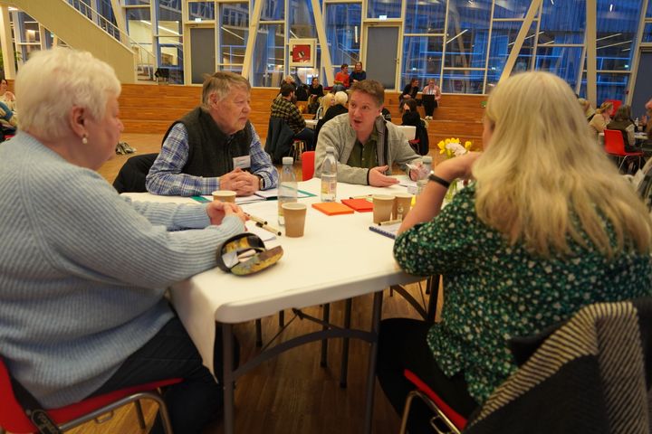 Fem mennesker sidder omkring et bord og snakker. I bagagrunden ses andre borde.