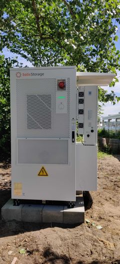 Et stort, hvidt kommercielt batterianlæg med teksten SolisStorage og et gult advarselsskilt foran. Anlægget er placeret udendørs på fliser i skyggen under grønne træer.
