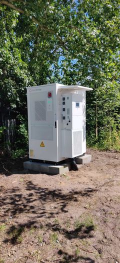 Et hvidt SolisStorage batterianlæg i profil, opstillet på et fundament af fliser udendørs på en grusplads omgivet af grønne buske og træer.